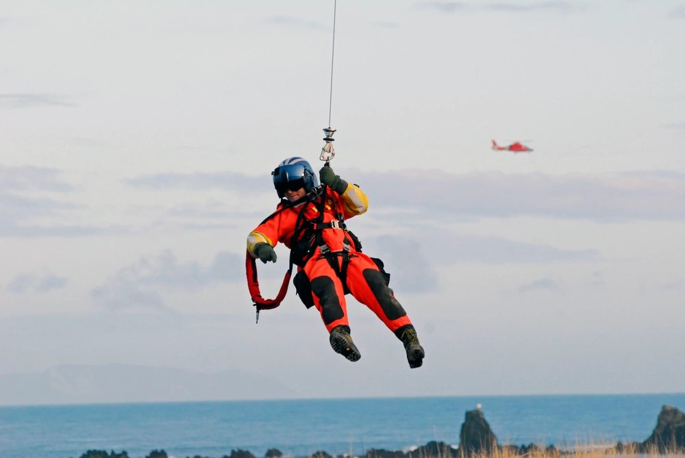 A search and rescue winchman in an orange dry suit hoisted on a helicopter cable above the sea, with a red rescue helicopter beyond